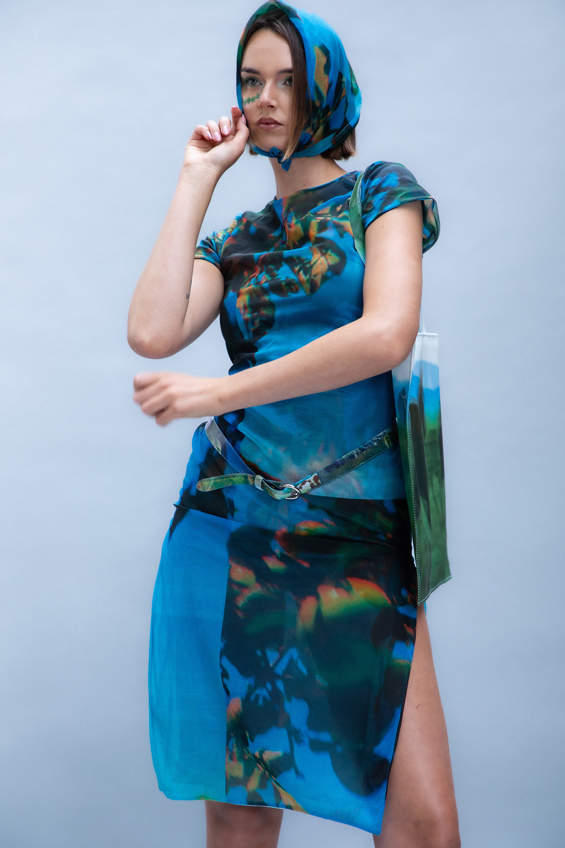 Model in a blue dress and matching headwrap posing with one are to their face against a gray backdrop